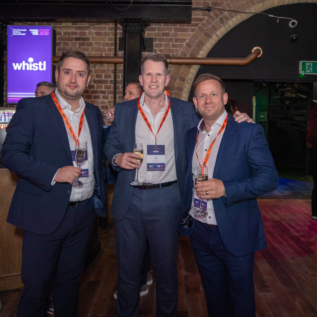 Three men in suits with event badges stand together, smiling and holding drinks at a networking event in a modern venue.