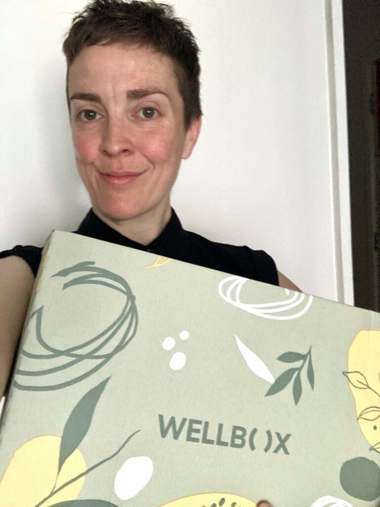 Woman smiling and holding a decorative WELLBO(X) box in front of a white wall.